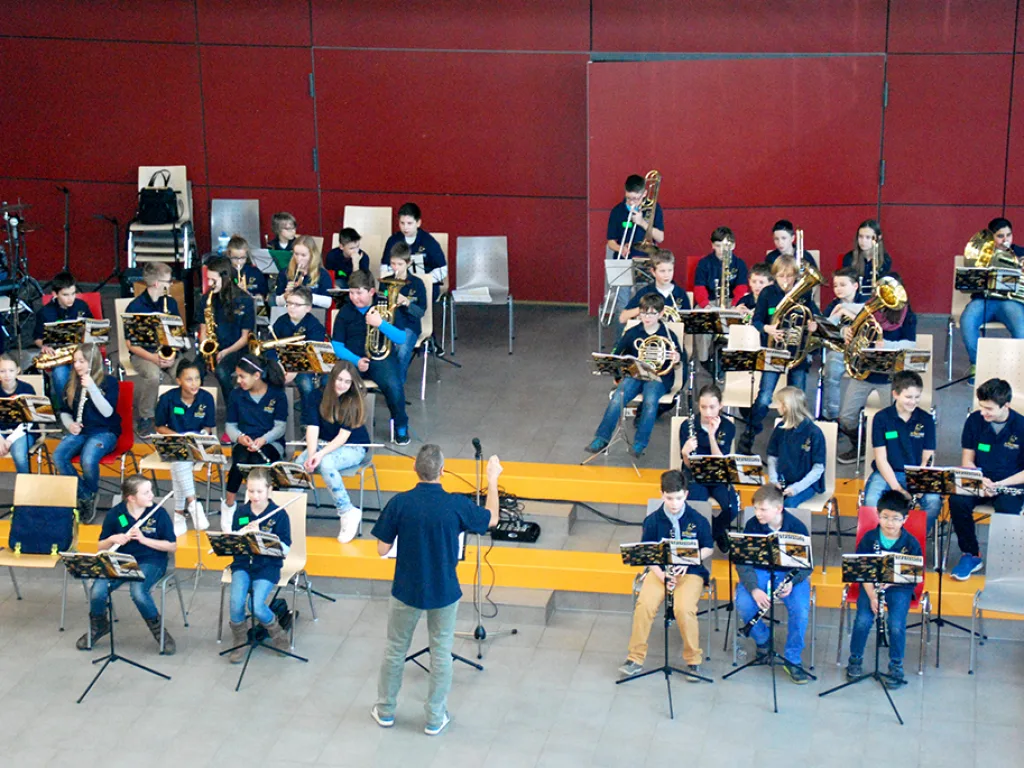 Das Blasorchester spielt vor der roten Wand, ein Dirigent leitet.
